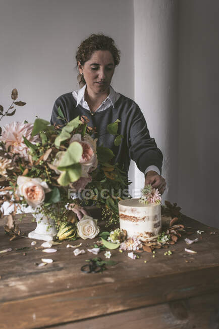 Vista lateral de dama colocando plato con sabroso pastel decorado brote de flor en la mesa de madera con racimo de crisantemos, rosas y ramitas de plantas en jarrón entre hojas secas sobre fondo gris - foto de stock