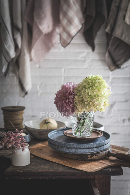 Деревянный стол со свежими розовыми хризантемами и белыми гортензиями в вазе между сковородкой и кухонной утварью рядом с посудой, висящей на твисте с булавками — стоковое фото