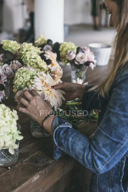 Senhora feliz perto de mesa de madeira com cachos de crisântemos frescos, rosas e galhos de plantas em vasos em fundo cinza — Fotografia de Stock
