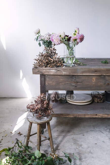 Mesa de madera con utensilios de cocina y ramos de flores frescas en jarrones con agua cerca de la pared blanca - foto de stock