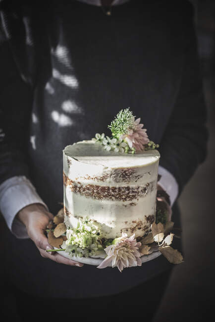 Вид збоку врожаю щаслива дама тримає тарілку зі смачним тортом, прикрашеним бутоном хризантем і сухим листям в кімнаті — стокове фото