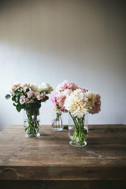 Mesa de madeira com utensílios de cozinha e buquês de flores frescas em vasos com água perto da parede branca — Fotografia de Stock
