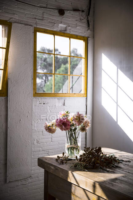 Mesa de madera con utensilios de cocina y ramos de flores frescas en jarrones con agua cerca de la pared blanca - foto de stock