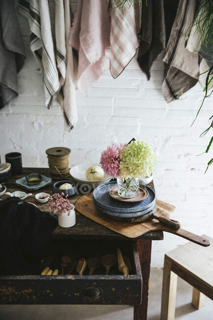 Wooden table with bunch of fresh pink chrysanthemums and white hydrangea in vase between frying pan and kitchenware near dish cloths hanging on twist with pins — Stock Photo