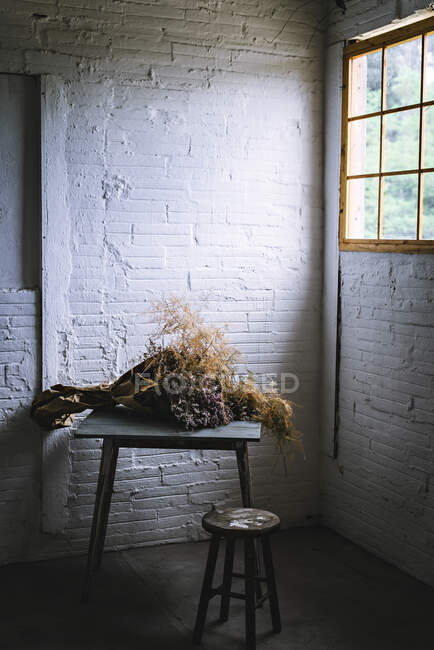 Concepto de ramo de ramitas de coníferas secas en papel artesanal sobre mesa cerca de taburete en habitación gris turbia con paredes de ladrillo - foto de stock