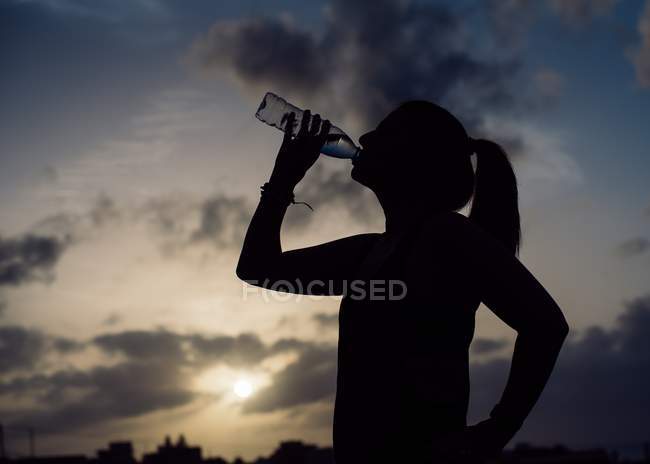 Silhouette of lady in sportswear drinking water from bottle on wonderful heaven background with clouds in evening — Stock Photo
