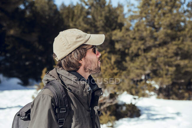 Side view of young man in sunglasses and cap with backpack looking away between winter forest in Cerdanya, France — Stock Photo
