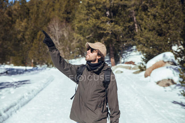 Seitenansicht eines jungen Mannes in Sonnenbrille und Mütze mit Rucksack, der zwischen Winterwald in Cerdanya, Frankreich, wegschaut — Stockfoto