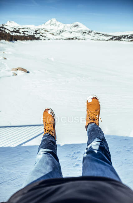 Обрезание мужских ног в джинсах и зимних сапогах, сидя на снегу в солнечный день возле холмов в Черданье, Франция — стоковое фото
