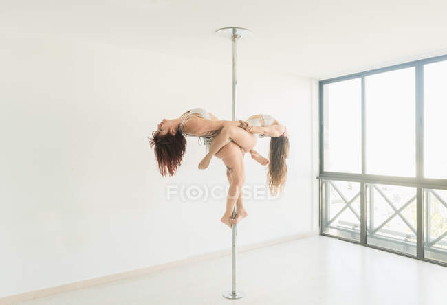 Side view of young slim ladies training on pole in light room near window — Stock Photo