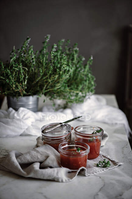 Pequeños frascos con deliciosos tomates frescos mermelada casera cerca de hierbas y servilleta en la mesa sobre fondo borroso - foto de stock