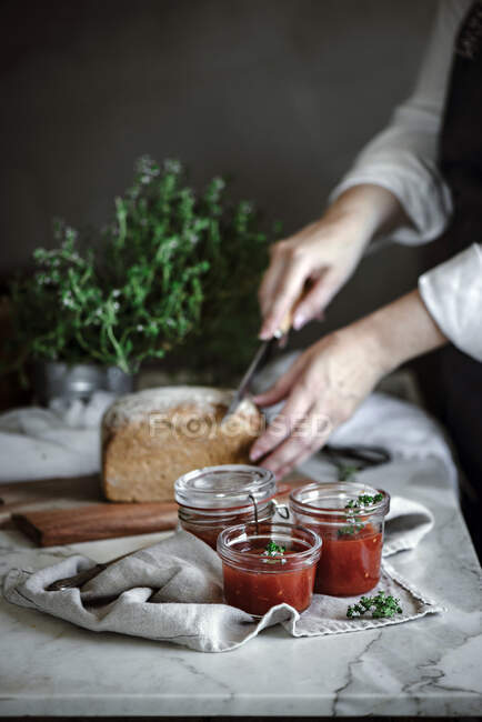Обрізані руки, що ріжуть хліб на серветці біля ножа та банок з помідорами домашнє варення на розмитому фоні — стокове фото