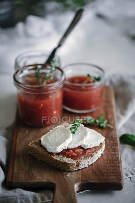 De arriba sándwich de rebanada de pan de centeno fresco con queso y hierba cerca de latas con tomates mermelada casera sobre tabla de cortar sobre fondo borroso - foto de stock