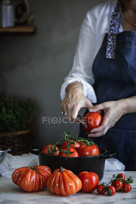 Coltivazione signora con coltello taglio verdure vicino pentola con pomodori freschi rossi sul tavolo — Foto stock