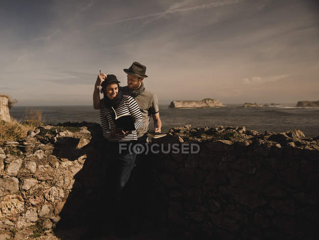 Стильный парень в шляпе и элегантная женщина в кепке читают тома у скал на берегу моря и чудесного неба — стоковое фото