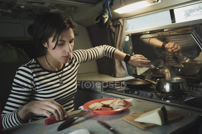 Woman at table with dish of food, cheese and cutlery taking sausage from pot on cooker in mobile home — Stock Photo
