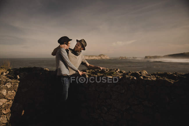 Vista lateral de elegante pareja elegante cerca de rocas en la costa cerca del mar y el cielo maravilloso - foto de stock