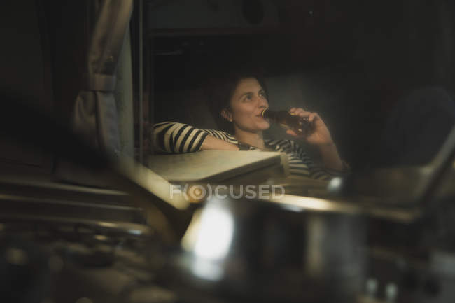 Reflection of young woman drinking from bottle and sitting on sofa near cooker — Stock Photo