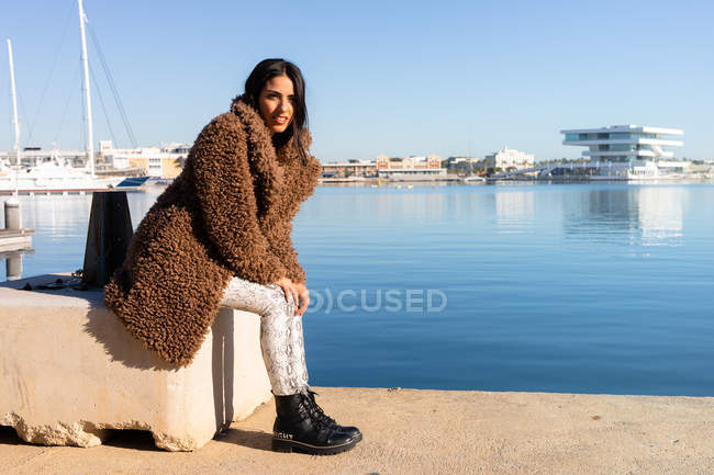 Vue latérale d'une jolie jeune femme en manteau tendance assise sur un bloc près de l'eau calme dans le port de la ville côtière — Photo de stock