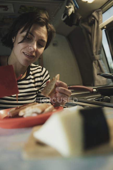 Mujer en la mesa con plato de comida, queso y cubiertos tomando salchicha de la olla en la cocina en casa móvil - foto de stock