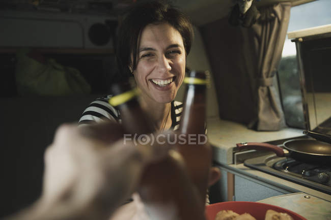 Junge brünette fröhliche Frau klirrt Flaschen mit Mann in der Nähe von Elektroherd in Mobilheim — Stockfoto