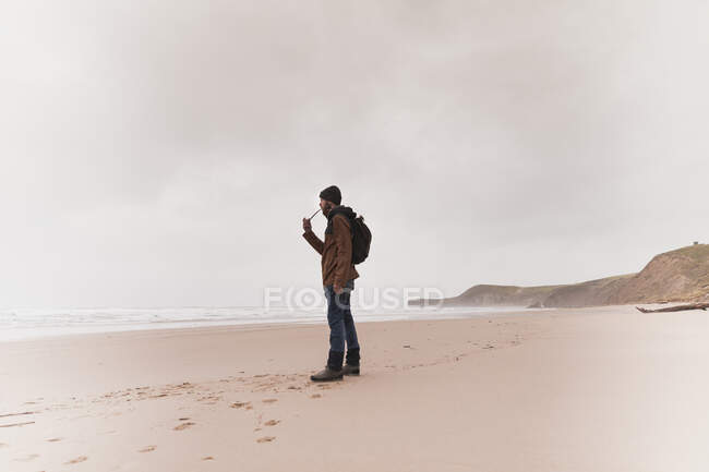 Вид сбоку парня в теплой одежде с рюкзаком курительной трубки на песчаном побережье у моря и небо в облаках — стоковое фото