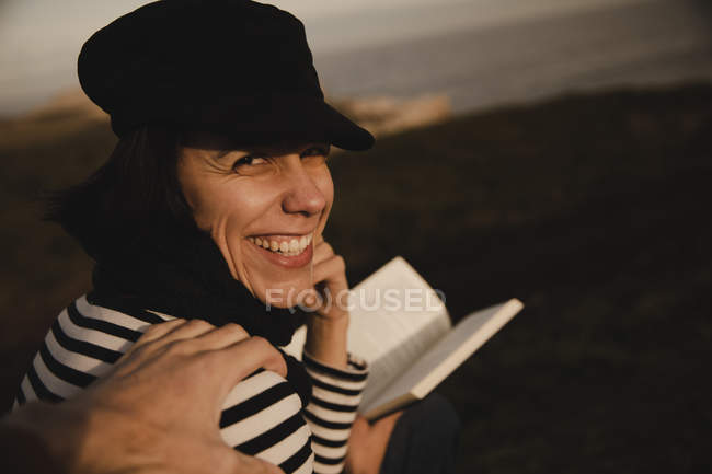Back view of cheerful elegant woman in cap holding volume with hand on shoulder on blurred background — Stock Photo