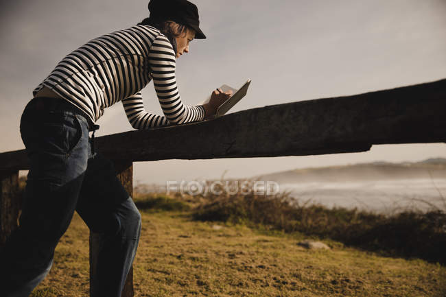 Elegant woman in cap taking notes in notepad and leaning on seat on coast near waving sea — Stock Photo