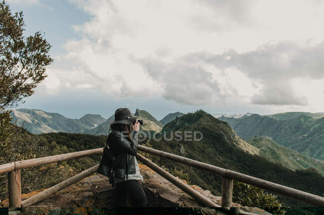 Side view of elegant lady in hat and leather jacket shooting on camera amazing view of peaks of hills and cloudy sky — Stock Photo
