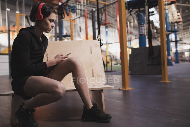 Side view of woman in sportswear with headphones and gadget listening music and sitting in gym — Stock Photo