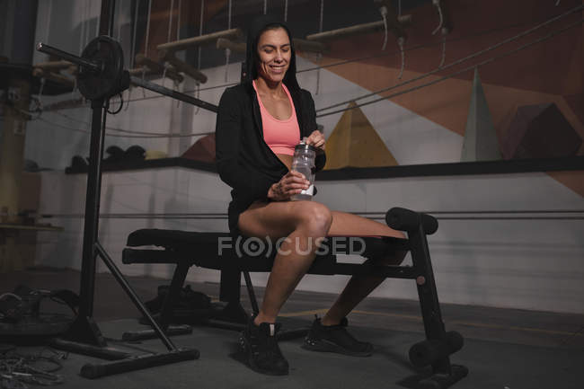 Junge schlanke, fröhliche Frau in Sportbekleidung mit Wasserflasche sitzt auf Bank im Fitnessstudio — Stockfoto