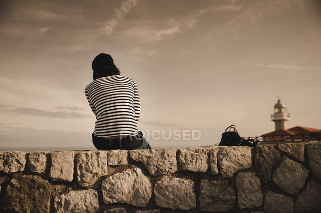 Вид сзади элегантной женщины в кепке, сидящей на лугу рядом с маяком на берегу и прекрасным небом — стоковое фото