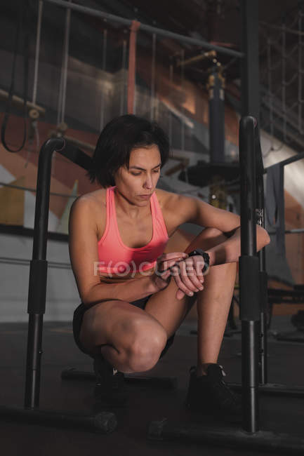Jeune femme sportive mince en vêtements de sport vérifier l'heure sur la montre et assis entre les barres parallèles dans la salle de gym — Photo de stock