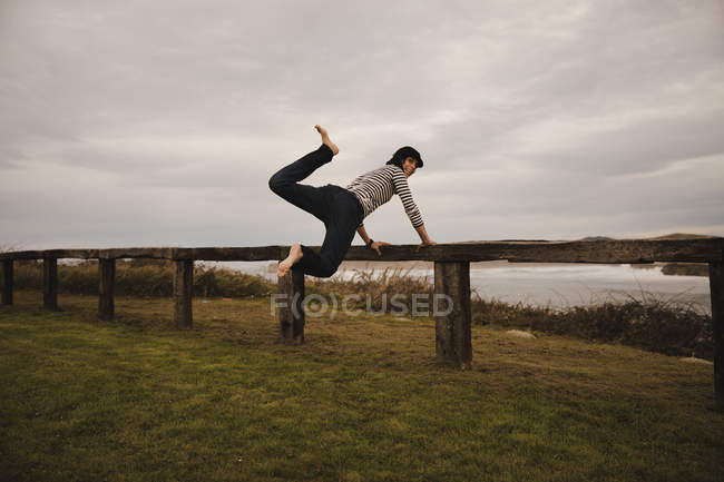 Вид сбоку на элегантную жизнерадостную женщину в кепке, прыгающую рядом с сиденьем на берегу рядом с морем и облачным небом — стоковое фото
