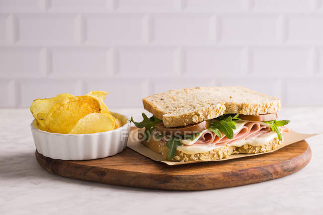 Вкусный сэндвич с ветчиной, сыром и зеленью на деревянной доске с картофельными чипсами на белом фоне — стоковое фото