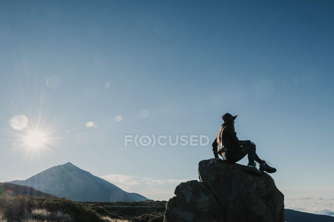 Вид збоку на невпізнавану мандрівну жінку, яка сидить на скелі на схилі пагорба в сонячний день — стокове фото