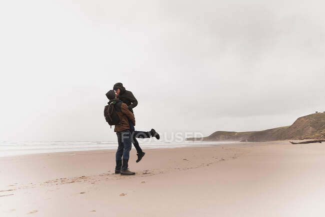 Вид збоку на молодого чоловіка з рюкзаком в теплому одязі, що тримається за руки і цілує жінку на піщаному пляжі біля моря і пагорбів — стокове фото
