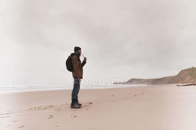 Вид збоку хлопця в теплому одязі з рюкзаком курильна труба на піщаному узбережжі біля моря і неба в хмарах — стокове фото