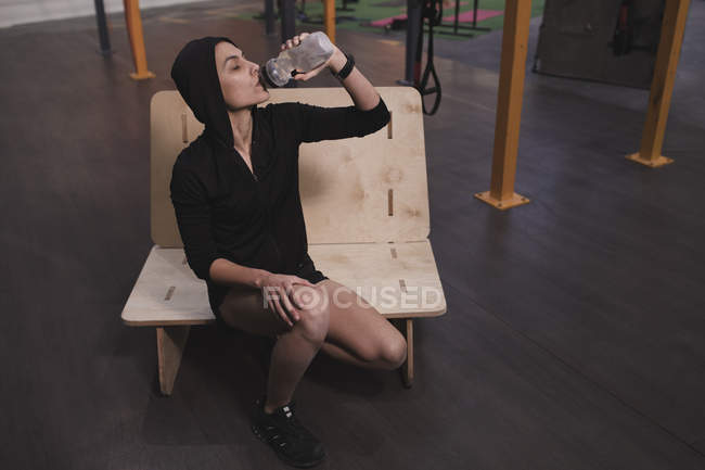 Mulher alegre em sportswear com garrafa de água sentado no banco no ginásio — Fotografia de Stock