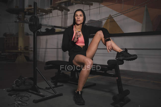 Mujer joven en ropa deportiva con barra de energía sentado en el banco en el gimnasio - foto de stock