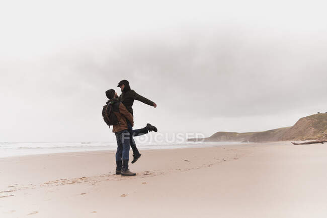 Side view of young man with backpack in warm wear holding on hands and kissing woman on sand beach near sea and hills — Stock Photo