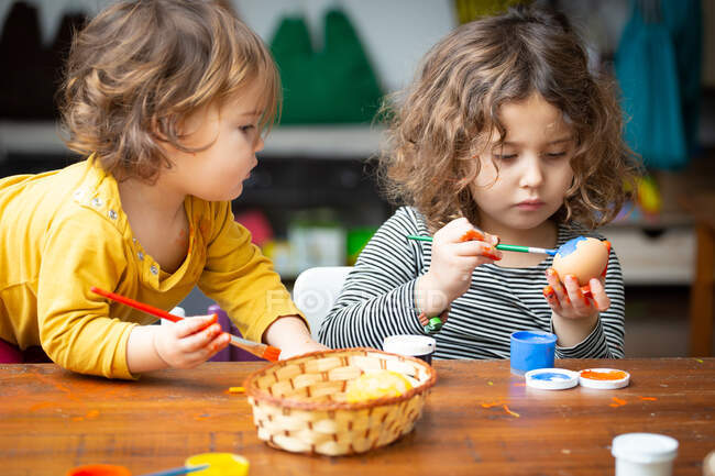 Two cute girls with brushes painting chicken eggs for Easter while sitting at wooden table together — Stock Photo