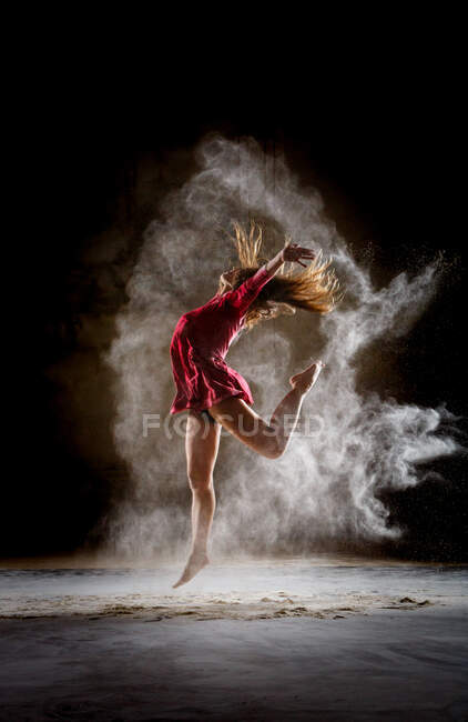 Seitenansicht der jungen schlanken Ballerina in rotem Kleid, die Bein und Hände zwischen Nebel im dunklen Raum bewegt — Stockfoto