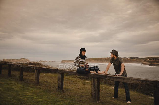 Junger Mann mit Hut neben eleganter Frau, die auf Handtrommel in Mütze auf Sitz in der Nähe der Küste von Meer und bewölktem Himmel spielt — Stockfoto
