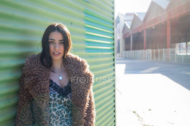Frau im trendigen Outfit blickt in die Kamera, während sie sich auf der Stadtstraße an eine grüne Wand lehnt — Stockfoto