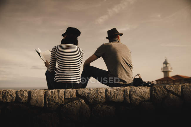 Vista laterale di elegante elegante coppia di lettura e seduta su rocce vicino faro e cielo meraviglioso — Foto stock