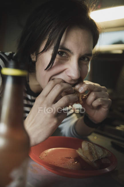 Mujer sentada en la mesa, mordiendo sándwich cerca de plato y botella de bebida y mirando a la cámara en casa móvil - foto de stock