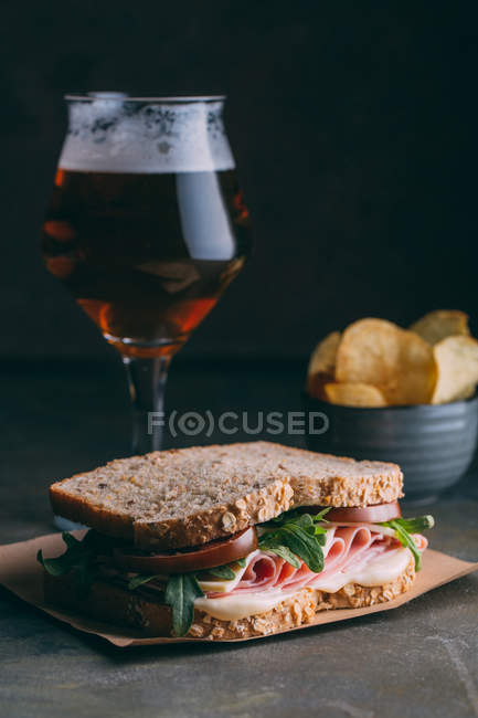 Delicious sandwich with ham, cheese and greens with glass of beer and chips on dark background — Stock Photo