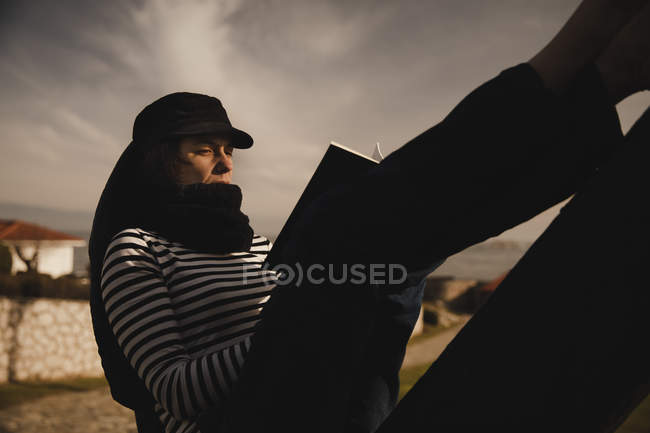 Вид сбоку элегантной женщины в кепке, читающей громко и сидящей на памятнике возле стройки на берегу моря — стоковое фото