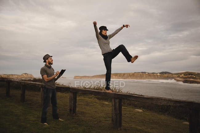 Jeune femme heureuse et homme en chapeaux tenant la main et s'amusant sur le siège sur la côte près de la mer agitant et ciel nuageux — Photo de stock